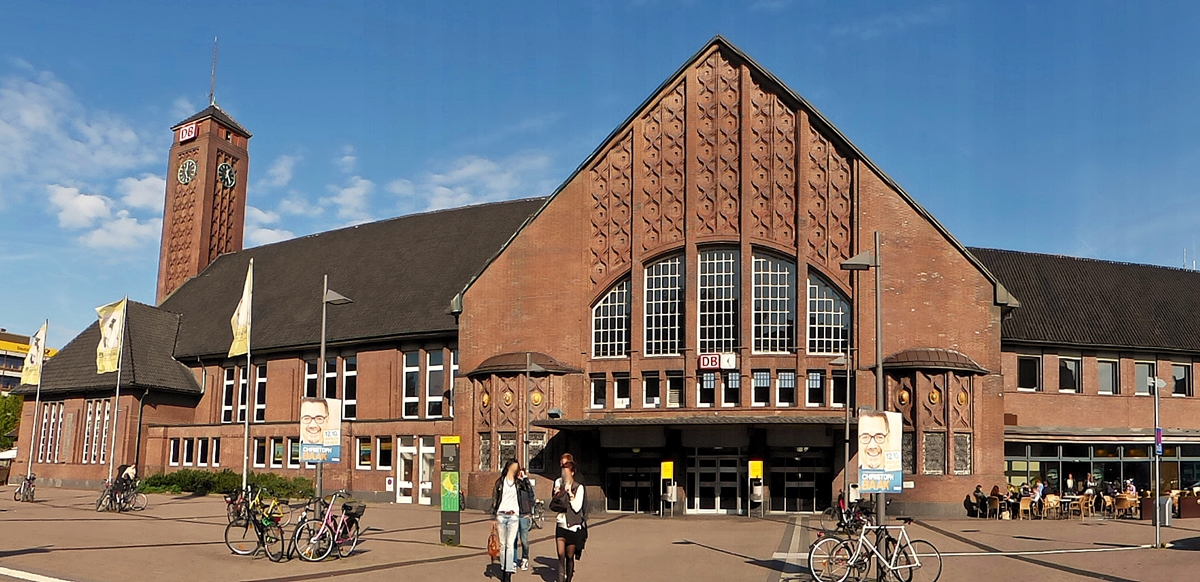 . Panorama Aufnahme von dem imposanten Empfangsgebudes von Oldenburg (Oldb) Hbf von der Straenseite aus aufgenommen. 11.10.2014 (Jeanny)

Der heutige Oldenburger Hauptbahnhof wurde nach vierjhriger Bauzeit am 3. August 1915 ohne groe Feierlichkeiten eingeweiht, nachdem er bereits am 1. August fr die ffentlichkeit zugnglich gemacht worden war. Der Groherzog besuchte den Bahnhof zusammen mit seinen Tchtern erst am 4. August. Das prunkvolle Gebude im Jugendstil wurde vom Architekten Friedrich Mettegang als Ersatz fr den alten Centralbahnhof gebaut. Fr den Groherzog von Oldenburg war ein separates Gebude, die sogenannte Frstenhalle, zum Besteigen der Zge vorgesehen. Im Zuge des Neubaues wurden die Gleise um rund 3,25 m angehoben. Das Gebude wurde bewusst an den Rand des Bahnhofes gelegt, da geplant war, den Bahnhof zu einem Durchgangsbahnhof umzufunktionieren. Bis zu diesem Zeitpunkt mussten Reisende, welche ber Oldenburg hinaus wollten, dort auch umsteigen. Das Hauptportal wurde in die Mittelachse des Hauptplatzes gelegt. Auf der rechten Seite des Gebudes wurden die Fahrkartenschalter und die Gepckabfertigung angeordnet, auf der linken Seite befanden sich die Wartesle fr die I. bis IV. Klasse. Neben dem Tunnel fr die Fahrgste gab es noch einen separaten Post- und Gepcktunnel mit Aufzgen zu den Bahnsteigen. Der Tarif fr die Bahnsteigkarten betrug zu diesem Zeitpunkt 30 Pfennige pro Person.

Am 21. April 1945 wurde der Hauptbahnhof bei einem Luftangriff auf Oldenburg schwer beschdigt.