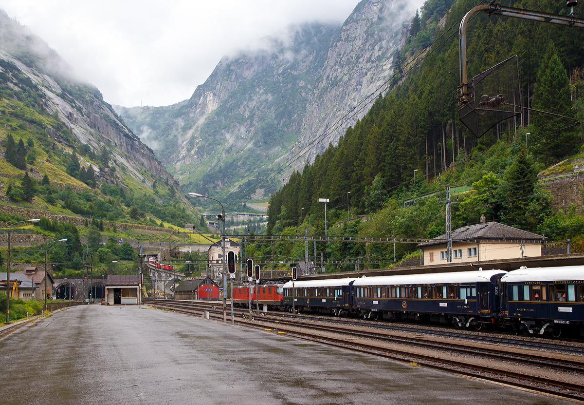 
Bahnhof Göschenen am 02.08.2019:
Während die SBB Re 8/8 (Re 4/4-11194 und Re 4/4-11156) mit dem Venice Simplon-Orient-Express (VSOE) nach Venedig im Bahnhof hält, schiebt eine MGB Deh 4/4 einen Schöllenenbahn-Zug über die Reuss in Richtung Andermatt.

Gut sichtbar (links im Bild die beiden Tunneleingänge), das Nordportal von dem über 100 Jahre alten und ca. 15 km langen Gotthardtunnel (italienisch Galleria del San Gottardo Gotthardtunnel).

Leider war es am regen als der VSOE hielt, aber man kann ja nicht alles verlangen.....