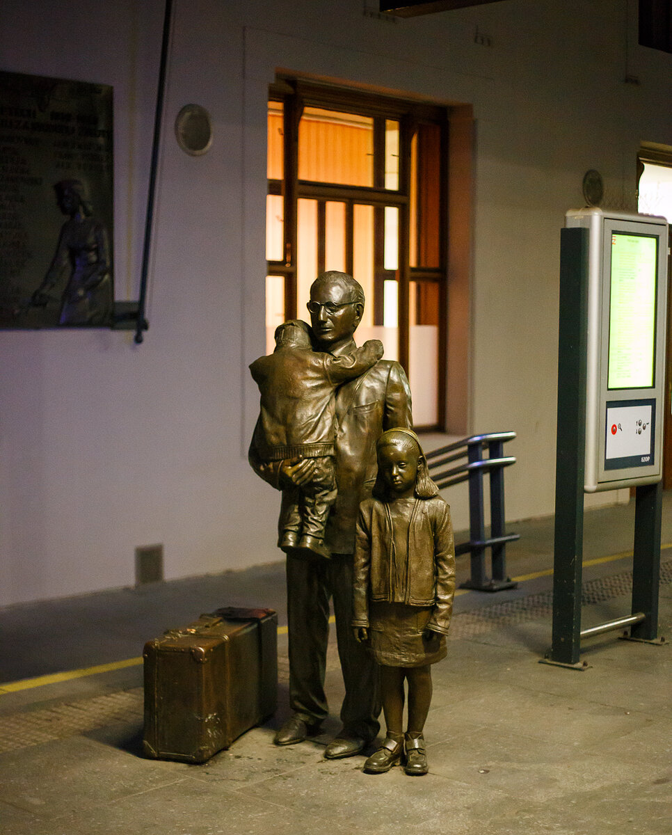 Bronzestatue einer tschechischen Familie am Bahnsteig 1 vom Prager Hauptbahnhof (Praha hlavni nadrazi) am 24.11.2022.  Die Skulpturengruppe ist von der Künstlerin Flor Kent. Ein Mann mit zwei Kinder und einem Koffer stehen in Bronze gegossen am Bahnsteig 1. Es sind sehr traurige Gesichter. Was ist die Geschichte dazu:

Das Denkmal ist sehr eindrücklich gestaltet. Die Personen stehen ohne Sockel quasi auf Augenhöhe mit den Wartenden am Bahnsteig 1 des Prager Hauptbahnhofs (Praha hlavní nádraží). Eine Tafel verrät Titel des Kunstwerks: Pro dítě (Für das Kind), Der Text dankt Sir Nicholas Winton (1909 – 2015), der kurz vor Beginn des Zweiten Weltkriegs die Rettung von 669 Kindern jüdischen Glaubens oder Herkunft aus der Tschechoslowakei vor dem Holocaust organisierte. Der Großteil davon waren Kinder aus Familien jüdischer Flüchtlinge aus Deutschland und Österreich, die in der Tschechoslowakei Asyl bekommen haben.
