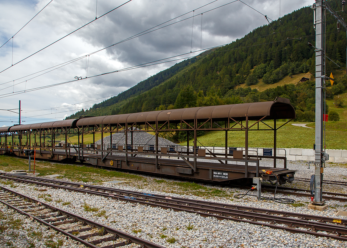 Der MGB Sklv 4811 Autotransportwagen Furkatunnel (Verladewagen) der Matterhorn-Gotthard Bahn, ex FO Sklv 4811 (Furka-Oberalp Bahn), der Serie 4811 bis 4827 für den Transport durch den Furka-Basistunnel abgestellt am 07.09.2021 beim Bahnhof Oberwald (1.365 m ü. M). 

Für die ROLLENDE STRASSE durch den Furka-Basistunnel beschaffte die Furka-Oberalp Bahn 1980 zwei komplette Zugkompositionen jeweils bestehend aus einer Zuglok Ge 4/4 III 81 oder 82, 2 Rampenwagen, 6 Transportwagen (Verladewagen) und 1 Steuerwagen. Seit der Wintersaison 1982 verkehren diese Züge mit hoher Auslastung. Die Gesamtlänge des Zuges beträgt 201 Meter. Um auch Busse und LKW durch den Tunnel transportieren zu können, wurde das entsprechende Wagenprofil der Vollspur von SBB bzw. BLS gewählt. Die maximale, nutzbare Höhe beträgt 4,50 Meter und die Breite 2,70 Meter.

Da die Verladungen auch im Umfahrtunnel Oberwald stattfinden, erhielten die Wagen seitliche Leuchtstoffröhren. Die Wagen sind für die im Tunnel geltende Höchstgeschwindigkeit von 90 Km/h ausgelegt.
Bei Ausfall einer Ge 4/4 III ist es möglich, die Kompositionen auch mit HGe 4/4 I, HGe 4/4 II, Deh 4/4 I oder Deh 4/4 II zu führen. Mitte der Neunzehnhundert-Achtziger Jahre wurde eine komplette Wagengarnitur nachbestellt. Nach Fusion der FO und der BVZ am 1.1.2003 zur Matterhorn-Gotthard Bahn sind alle Fahrzeuge auf die MGB umgezeichnet worden.

TECHNISCHE DATEN:
Vierachsiger Verladewagen (Autotransporter)
FO, Serie Nr. Sklv 4811 - 4822
Anzahl:  12
Baujahr:  1980
Hersteller:  SWS Schieren
Länge über Puffer: 20.900 mm
Gewicht:  41,4 t
Ladegewicht:  20 t
Ladefläche:  53 m²
Maximale Ladehöhe:  3.400 mm
Breite des Wagenkastens:  2.900 mm
Höchstgeschwindigkeit:  90 km/h
Bremsen:  Oe, Laü, mFB, VL

Gezogen werden die Wagen von der eigens für diese Zugkompositionen angeschafften neuen Lokomotiven Ge 4/4 III mit den Nummern 81 und 82, einer Weiterentwicklung der RhB Ge 4/4 II. Eine Zugkomposition sieht folgendermaßen aus:
An der Spitze des Zuges befindet sich immer auf der Seite Realp die Lokomotive. Es folgt ein Rampenwagen (Auffahrwagen) Sklv 4801 bis 4807, dann sechs Verladewagen (Sklv 4811 bis 4827), wieder ein Rampenwagen und als Abschluss ein Steuerwagen (BDt 4361bis 4363) auf der Seite Oberwald. Die Rampen- und Verladewagen sind mit Kuppelstangen fest zu einem Blockzug verbunden. Die Rampenwagen wiederum sind gegen die Lok und den Steuerwagen mit einer automatischen Kupplung +GF+, Typ Brünig, ausgerüstet.

Der Furka-Basistunnel wurde trotz der vielen Querelen im Vorfeld des Baus und der immens hohen Baukosten von 300 Millionen Schweizer Franken ein voller Erfolg. Bis zu Eröffnung des Tunnels fehlte eine Winterverbindung. Jedes Jahr musste die Steffenbachbrücke auf der FO-Strecke im Herbst abgebaut und im Frühjahr wieder aufgebaut werden. Das Goms war in diesen 8 Monaten, solange war der Verkehr der FO vor dem Tunnelbau unterbrochen, mehr oder weniger vom Leben abgeschnitten. Es drohte die Entvölkerung, da auch geeignete Arbeitsplätze fehlten. In den Wintermonaten versah die Furka-Oberalp Bahn hauptsächlich Pflichtaufgaben, die fast nur noch sozialen Charakter hatten. Seitdem der Tunnel in Betrieb ist, hat sich die Situation grundlegend geändert. Das Goms und das ganze Wallis sind besser erreichbar, und der Wintertourismus hat Einzug gehalten. Generell wirkte sich die Eröffnung des Furka-Basistunnel positiv auf alle von der Furka-Oberalp Bahn bedienten Gebiete aus. Und die Autransport-Züge sind nicht mehr wegzudenken. Bis zu 180.000 Autos jährlich befördern die beiden Autotransportzüge durch den Furka-Basiatunnel.

Technische Daten Furka-Basistunnel:
Länge Oberwald bis Realp:  15,407 km
Maximale Steigung: 17,5 Promille
Höhendifferenz Portal Oberwald - Portal Realp:  160 m
Kreuzungsstellen im Tunnel:  2
Nutzlängen der Kreuzungsstelle:  je 500 m
Bauzeit:  1973 - 1982
Vorgegebene Durchfahrtsgeschwindigkeit: 55 - 75 km/h
Maximale Durchfahrtsgeschwindigkeit:  90 km/h
Fahrzeit:  15 - 22 Minuten

Der Furkatunnel ist 15,4 Kilometer lang und verbindet die Stationen Realp und Oberwald. Die einspurige Tunnelstrecke wird durch zwei je 774 Meter lange Tunnel-Ausweichen unterteilt. Der automatische Block erlaubt eine Zugfolge von 8 bis 10 Minuten.
