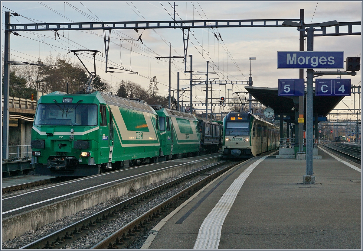 Die beiden BAM MBC Ge 4/4 22 und 21  schieben ihren bereits umfahrenen Güterzug mit 28 Achsen auf die Rollbockanlage, wo die Normalspurgüterwagen de BAM /MBC wieder ihr Normalspurgleis erreichte, Der Zug wird später mit der BAM MBC Re 4/4 II nach Gland gefahren. Rechts im Bild wartet ein BAM MBC Be 4/4  SURF auf die Abfahrt nach Bière.
Ganz rechts im Bild aber kaum erkennbar wartet der Gegenzug auf den Spur und Fahrrichtungswechsel; auf jedenfall ergibt die Kreuzung der beiden Züge eine  Kaisermanöver  vom Feinsten. 
22. Feb. 2017