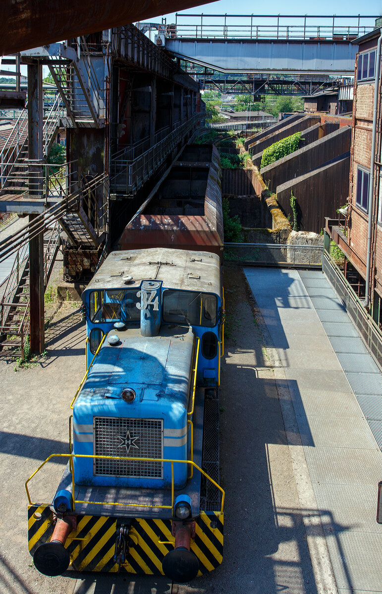 Die Dieselhydraulische Normalspur-Lokomotive Nr. 34, eine Henschel DHG 240 B  Esslinger , der Völklinger Hütte (ehem. Röchling'schen Eisen- und Stahlwerke GmbH in Völklingen/Saar, ab 1982 ARBED-Saarstahl GmbH, ab 1986 Saarstahl Völklingen GmbH) hier am 01 Juli 2025 abgestellt als Denkmal, mit zwei selbstentladende Schüttgutwagen der Gattung Fad oder Falns am Eingang der Rohstoffentladebunker in Richtung Hüttenbahnhof. Die Darstellung steht für den Rohstofftransport der Hütte zur Herstellung des Sintergutes. Die Lok war auch auf Bundesbahn Gleisen zugelassen. Heutiger Eigentümer der Lok und Wagen ist die Initiative Völklinger Hütte e.V..

Die zweiachsige dieselhydraulische Lok mit Antrieb über Gelenkwellen wurde 1969 von Henschel in Kassel unter der Fabriknummer 31210 gebaut und an die Röchling'sche Eisen- und Stahlwerke GmbH in Völklingen als Lok 34 geliefert. Weiterer Lebenslauf/Umfirmierungen: 
01.01.1971 > Stahlwerke Röchling-Burbach GmbH, Völklingen  34  
Juni 1982 	 > ARBED-Saarstahl GmbH, Völklingen  34  
Juni 1986 	 > Saarstahl Völklingen GmbH, Völklingen  34  
10.05.1989 > Saarstahl AG, Völklingen  34  
Später an MGS - Metallurgische Gesellschaft Saar mbH, Völklingen  34  
März 1999 	an Weltkulturerbe Völklinger Hütte - Europäisches Zentrum für Kunst und Industriekultur GmbH, Völklingen und Aufstellung als Denkmal. 

Die Lokomotive war bis zu einem Motorschaden 1997 in verschiedenen Bereichen eingesetzt: Transport von Duplexeisen zwischen der Mischerhalle und dem Siemens-Martin-Werk, im Bereich des Siemens-Martin-Werks, der Elektroöfen und des Hammerwerks sowie nach Einbau einer Funkfernsteuerung im Hafenbereich der Saarstahl AG.