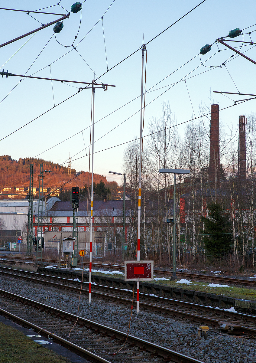 
Niederschelderhütte (zu Mudersbach, DB mäßig nur Niederschelden) den 13.02.2018:Die Oberleitung ist geerdet und stromlos....

Die Erdungsstangen sind ein gehangen, am Haken oberen Ende der Stange ist das Kabel angeschlossen, welches mittels Schienenklemme an das Gleis angeschlossen ist und somit ist die Oberleitung geerdet. An der vorderen Stange ist zudem eine Sh 2 - (Schutz-)Halttafel befestigt. Diese Tafel gebietet Halt und ersetzt ein haltzeigendes Hauptsignal. Nachts leuchtet an der Tafel zusätzlich ein rotes Licht.

Eine Erdungsstange, auch Erdungsstab, ist eine isolierende Stange mit deren Hilfe die Oberleitung von Hand geerdet werden kann. Am oberen Ende ist ein Haken fürs Erden fest angebracht. Das daran befindliche Kupferkabel wird dann an die Schiene (Gleis) an geklemmt, um so den Erdschluss herstellen. Mit der Erdungsstange wird im Rahmen von Installations- oder Wartungsarbeiten an freigeschalteten Oberleitungen ein Kurzschluss der Fahrleitung hergestellt, um Stromunfälle durch elektrische Spannungen zu verhindern.

Die Erdungsstange ist eine rote/weiße (oder gelbe) Stange aus glasfaserverstärkten Kunststoff (GFK). Je nach Einsatzzweck kann die Stange als Teleskopstange ausgeführt sein oder aus mehreren Einzelteilen zusammensteckbar sein. Sie ist so leichter zu transportieren und vor der Witterung geschützt in Taschen unterzubringen. Durch das Zusammenstecken aus einzelnen Elementen werden Längen von mehreren Metern erreicht. Die Erdungsstange ist dabei auf eine solche Länge zusammenzubauen, dass das zu erdende Anlagenteil erreicht wird und Sicherheitsabstände nicht unterschritten werden.

Die korrekte Vorgehensweise beim Erden mit Erdungsstangen ist eine angepasste Abfolge wie bei den Fünf Sicherheitsregeln:
1. Freischalten der Fahrleitung. (Oberleitung ausgeschaltet)
2. Gegen Wiedereinschalten sichern
3. Spannungsfreiheit feststellen: (Teilweise haben Erdungsstangen auch Spannungsprüfer eingebaut)
4. Erdungskabel Schienenfuß befestigen
5. Erden und kurzschließen

So wird sichergestellt, dass Personen nicht durch eventuell auftretende Kriechspannungen oder gar durch das unbeabsichtigte Einschalten der Fahrleitung gefährdet werden.
