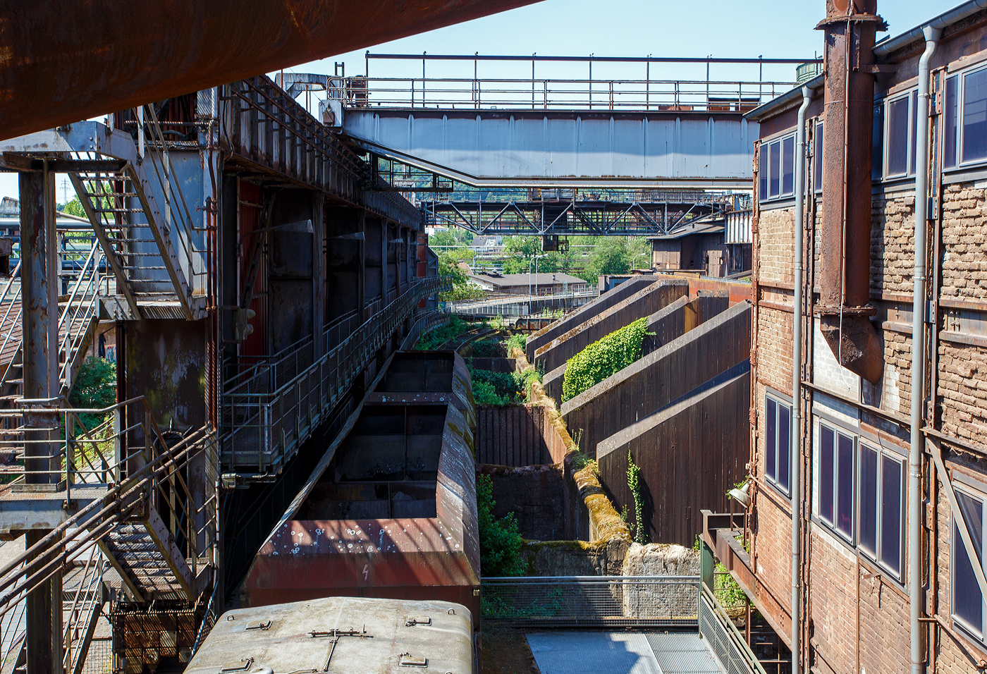 Am Tiefbunker (Rohstoffentladebunker der Sinterhalle) der Völklinger Hütte (ehem. Röchling'schen Eisen- und Stahlwerke GmbH in Völklingen/Saar, ab 1982 ARBED-Saarstahl GmbH, ab 1986 Saarstahl Völklingen GmbH) hinter der Normalspur-Denkmallok 34 stehen zwei vierachsige offene Schüttgutwagen (Sattelwagen) mit schlagartiger Schwerkraftentladung, hydraulischem Klappenverschlusssystem vermutlich der Gattung Falns (genaue Bezeichnungen kann man nicht erkennen), hier am 01 Juli 2025. Die Darstellung steht für den Rohstofftransport der Hütte zur Herstellung des Sintergutes. Die Lok war auch auf Bundesbahn Gleisen zugelassen. Heutiger Eigentümer der Lok und Wagen ist die Initiative Völklinger Hütte e.V..