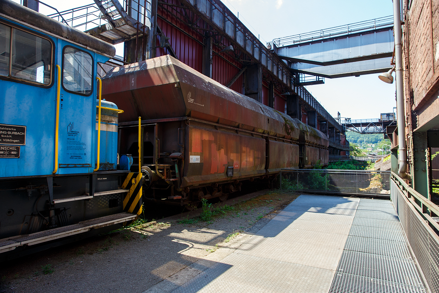 Am Tiefbunker (Rohstoffentladebunker der Sinterhalle) der Völklinger Hütte (ehem. Röchling'schen Eisen- und Stahlwerke GmbH in Völklingen/Saar, ab 1982 ARBED-Saarstahl GmbH, ab 1986 Saarstahl Völklingen GmbH) hinter der Normalspur-Denkmallok 34 stehen zwei vierachsige offene Schüttgutwagen (Sattelwagen) mit schlagartiger Schwerkraftentladung, hydraulischem Klappenverschlusssystem vermutlich der Gattung Falns (genaue Bezeichnungen kann man nicht erkennen), hier am 01 Juli 2025. Die Darstellung steht für den Rohstofftransport der Hütte zur Herstellung des Sintergutes. Die Lok war auch auf Bundesbahn Gleisen zugelassen. Heutiger Eigentümer der Lok und Wagen ist die Initiative Völklinger Hütte e.V..