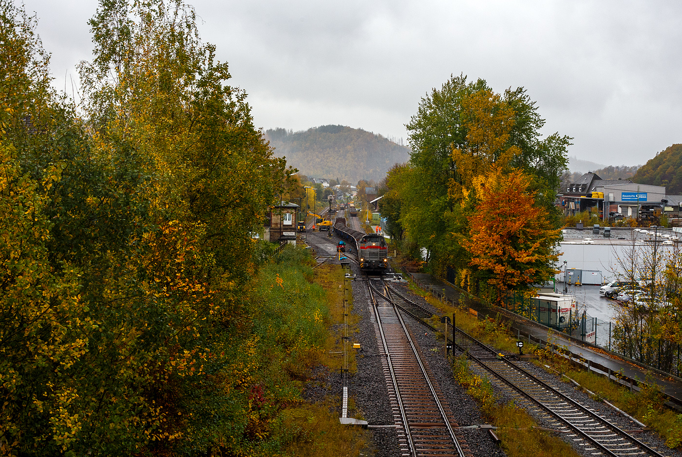 Baustelle Bahnhof Herdorf am 25 Oktober 2025, Blick von der Br�cke Wolfsweg („Achenbachs Br�cke“) Blickrichtung Betzdorf, es sch�ttete ohne Unterlass, dazu noch heftige Windb�en, aber die Arbeiten zur Erneuerung der Weiche 3 und der R�ckbau mit L�ckenschluss f�r die drei aufeinander folgenden Weichen 22, 23 und 24 (Erneuerung Anschluss von Weiche 26 an Gleis 4) haben begonnen. So musste ich doch dem widrigen Wetter an die Strecke.

Links ist das alte Gleis und Weichen demontiert und das Gleisbett vorbereitet.

Vorne steht die Vossloh G 12 – 4120 001-7 „Karl August“ (92 80 4120 001-7 D-KAF) der KAF Falkenhahn Bau AG mit einem Schotterzug. Hinten ist die 271 022-6 „Charlotte“ (92 80 1271 022-6 D-KAF) eine Vossloh/MaK G 1000 BB der KAF Falkenhahn Bau AG. ex MRCE bzw. ex HGK DH 49, mit einigen Flachwagen.

Bei dem Wetter haben die Gleisbauarbeiter mein vollstes Mitgef�hl. Da haben es die Lokf�hrer und Baggerfahrer durch den Schutz ihrer Kabine schon besser. 