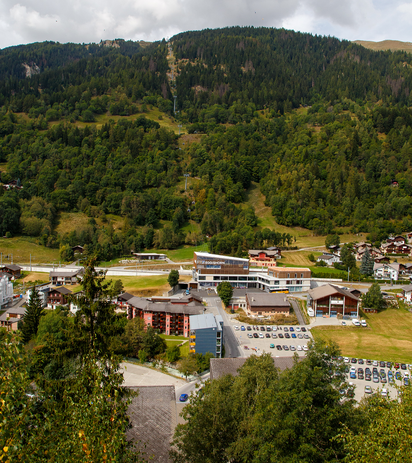 Blick auf den neuen V-Hub Fiesch am 07 September 2021 aus einem MGB-Zug. Ein Jahrhundertbau in Fiesch, MGB Bahnhof (1.063 m .M.) und Bus-Terminal mit dem direkten Zugang zur Talstation der neuen 10er-Kabinenbahn der Doppelmayr/Garaventa-Gruppe hinauf auf die Fiescheralp. 

Die neue 10er-Gondelbahn Fiesch - Fiescheralp / Khboden, Baujahr 2019 von der Doppelmayr/Garaventa-Gruppe ist mit 7 m/s Fahrgeschwindigkeit die schnellste 10er-Gondelbahn der Schweiz. Sie ist eine sogenannte D-Line-Bahn der Doppelmayr/Garaventa-Gruppe mit Seillageberwachung RPD. Als Betriebsmittel kommen die neuen CWA Kabinen Omega V zum Einsatz. Als Antrieb wurde der 867 kW starke Doppelmayr Sector Drive (DSD) verwendet, ein permanenterregter Synchronmotor, der mit modernster Frequenzumrichtertechnologie arbeitet und sich auch fr steile Anlagen wie die hier in Fiesch sehr gut eignet.

Die neue Gondelbahn ersetzt zwei Pendelbahnen von Fiesch zur Fiescheralp, einmal mit 100er-Kabinen und einmal mit 34er-Kabinen. Die Talstation der hochmodernen Anlage befindet sich im neuen V-Hub (Knotenpunkt ffentlicher Verkehr) in Fiesch, wo direkter Anschluss an die Matterhorn-Gotthard-Bahn und das Postautonetz besteht.

Ein Ausbau der Bahn mit einer zweiten Sektion vom Khboden Richtung Eggishorn als Ersatz fr die Luftseilbahn Fiesch-Eggishorn ist angedacht und bereits genehmigt.

Immer mehr Skitouristen nutzen den ffentlichen Verkehr fr die Fahrt in den Wintersport. Voll diesem Trend entsprechend haben die Aletsch Bahnen, die Matterhorn-Gotthard-Bahn (MGB) und die Gemeinde Fiesch als Bauherren mit dem Architekten und Investor Hans Ritz die neue V-Drehscheibe in Fiesch als Gemeinschaftsprojekt gebaut. Der neue Verkehrsknotenpunkt besteht aus der Talstation der neuen 10er-Kabinenbahn auf die Fiescheralp, einem komplett neuen Bahnhof der MGB und einem Postbus-Terminal. Durch die Integration und Verknpfung aller Verkehrstrger an einem Ort wurden kurze, schnelle und barrierefreie Umsteigwege geschaffen.

Fr das Projekt hat die MGB den Haltepunkt fr ihre Zge vom bisherigen Bahnhofsgebude 400 m nach Norden verlegt und in den V-Hub integriert. Dadurch ist ein komplett neuer Bahnhof mit zwei Auenbahnsteigen sowie einem 600 m langen neuen Doppelgleis fr eine optimierte Kreuzungsmglichkeit der Zge entstanden. Eine besondere Herausforderung bei der Gleisverlegung stellte die 36 m lange Bogenweiche mit einem Gewicht von 35 t dar. Sie wurde in drei Teilen angeliefert und dann mit einem Kran ber die Dcher gehievt und eingebaut.

Die neue Kabinenbahn ersetzte die 100er-Pendelbahn Fiesch – Fiescheralp (Khboden) von Garaventa aus dem Jahr 1974. Fr die neue Bergstation wurde die bestehende Bausubstanz der abgebauten Pendelbahn erweitert und fr die Garagierung der Kabinen verwendet. Bei der Talstation wurden 760 m“ und bei der Bergstation 1.100 m Kunststoff-Folie als Fassaden-verkleidung eingesetzt. Die Verkehrswege in den Stationen sind barrierefrei angelegt. Die Bahn ist mit der neuesten Kabinengeneration der Omega V von CWA ausgestattet, fr den Gtertransport stehen zwei Transportkabinen zur Verfgung.

Der V-Hub Fiesch wurde nach einer uerst kurzen Bauzeit von 14 Monaten am 7. Dezember 2019 feierlich erffnet. Mit rund 24 Mio. CHF bernahm die Aletsch Bahnen AG den Hauptanteil dieser Investition. Die MGB beteiligte sich mit 17,6 Mio. CHF, der Architekt Hans Ritz investierte 6 Mio. Das Vorzeigeprojekt V-Hub Fiesch macht eindrucksvoll deutlich, was durch Kooperation von verschiedenen Partnern erreicht werden kann.

TECHNISCHE DATEN der Seilbahn:
Hersteller:  Garaventa  (Doppelmayr/Garaventa-Gruppe)
Typ: 10er Gondelbahn (Ein-Seil-Umlaufbahn)
Baujahr: 2019
Hhe der Talstation:1.083 m . M.
Hhe der Bergstation: 2.227 m . M.
Hhendifferenz: 1.144 m
Abstand zwischen Tal- und Bergstation: 2.698 m
Anzahl der Sttzen: 15
Frderseildurchmesser: 56 mm
Antriebsstation: Berg (Spanneinrichtung Tal)
Motorleistung: 867 kW (im Betrieb) / 1.124 kW (beim Anfahren)
Geschwindigkeit:  7 Meter pro Sekunde
Effektive Fahrzeit: 7:48 Minuten
Frderleistung: 1.880 Personen pro Stunde
Kabinenanzahl: 48 und 2 Transport-/Gterkabinen
Kabinentyp: CWA Omega V