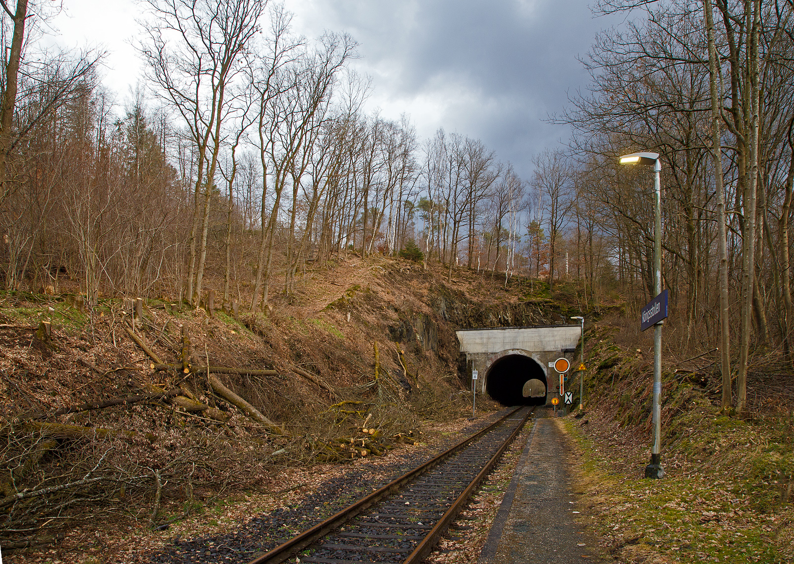 Der 137 m lange Herdorfer Tunnel hier am 18.03.2023 vom Hp Knigsstollen gesehen. 

Davor das Vorsignal (frs Einfahrtsignal Bf Herdorf) Vr 0 - Halt erwarten! Die gelbe Scheibe mit weiem Rand und gelbe Pfeil zeigt nach unten. Nachts leuchten auch die beiden gelben Lampen. Vor dem Signal die Ne 2 – Vorsignaltafel. Die Vorsignaltafel zeigt dem Lokfhrer, da das Signal, vor dem sie steht, Vorsignalfunktion hat, d.h., den Begriff eines nachfolgenden Hauptsignales vorankndigt. 
