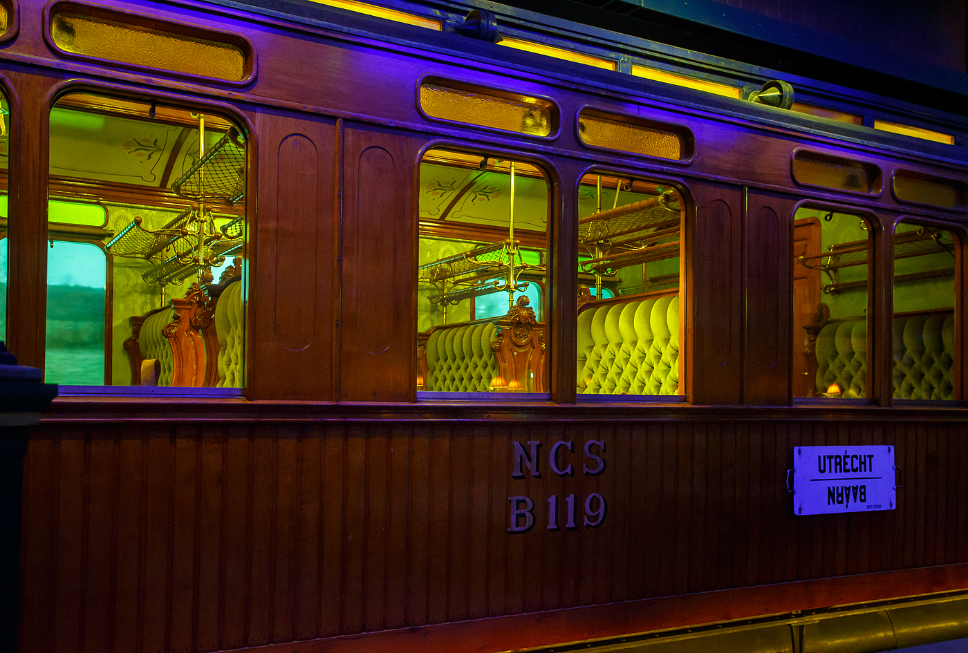 Detailbild des zweite Klasse Reisezugwagen NCS B 119, ein luxuriöser Teakholzwagen der ehemaligen NCS - Nederlandsche Centraal Spoorwegmaatschappij (Niederländische Zentralbahngesellschaft) am 24. Juni 2025 im Het Spoorwegmuseum (bis 2005 NSM - Nederlands Spoorwegmuseum / Niederländischen Eisenbahnmuseum) in Utrecht im ehemaligen Bahnhof Maliebaan. Nicht nur die Compagnie Internationale des Wagons-Lits (CIWL) für ihre Luxuszüg, wie beispielsweise dem Orient-Express, hatte solche Wagen.

Dieser äußerst luxuriösen Abteilwagen vom Typ BL8 (Serie B 111 – 127) wurde 1904 in der eigenen Werkstatt der NCS in Utrecht (Werkplaats NCS) gebaut. 

Bedingt durch den Ersten Weltkrieg wurde 1917 eine Zweckgemeinschaft der unabhängigen Eisenbahngesellschaften HSM beziehungsweise HIJSM (Hollandsche IJzeren Spoorweg-Maatschappij) und SS (Maatschappij tot Exploitatie van Staatsspoorwegen) gegründet. Da die NCS unter der Kontrolle der SS stand, wurde sie von diesem Moment an auch Teil der neuen Partnerschaft. Der Betrieb wurde am 1. Juni 1919 an die SS übertragen. Die beiden Gesellschaften errichteten einen gemeinsamen Bahnbetrieb, blieben aber rechtlich unabhängig. Dies änderte sich 1938, als HIJSM und SS vollständig fusionierten und in der AG NS (Nederlandse Spoorwegen, früher Nederlandsche Spoorwegen geschrieben) aufgingen. Das Rollmaterial wurde bereits 1921 zusammengelegt. Somit wurde ab 1924 der Wagen als NS B 1814 geführt, 1952 wurde er außer Dienst gestellt und für das Eisenbahnmuseum erhalten. Er kam 1977 vollständig restauriert als NCS B 119 dort an. Seitdem ist ein Paradewagen des Museums.

Anfang des 20. Jahrhundert war es den lokalen Eisenbahnen in den Niederladen gesetzlich verboten Waggons der 1. Klasse einzusetzen. Um doch wohlhabende Passagiere von Zeist und Baarn (einschließlich des Direktors der NCS) in passender Art zu befördern umging die NCS mit diesen als 2. Klasse Wagen klassifizierten äußerst luxuriösen Standard. Neben den teakholzgetäfelten Außenwänden ist auch der Innenraum opulent und gepflegt. Zu beiden Seiten des Mittelgangs befinden sich gepolsterte Doppelbänke mit Wendekissen. Im Winter war die plüschige Seite oben, im Sommer die kühlere, lederbezogene Seite. Lampen, Gepäckablagen, Aschenbecher, alles ist wunderschön verarbeitet und aus Kupfer gefertigt. Die Waggons wurden unter anderem auf den (Lokal-)Bahnen Utrecht–Den Dolder–Baarn und Utrecht–Bilthoven–Zeist eingesetzt. Insgesamt gab es siebzehn dieser Wagen.
