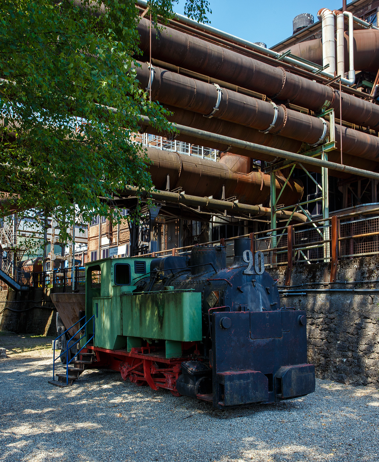 Die 785 mm Schmalspur-Dampflokomotive Nr. 20 der V�lklinger H�tte (ehem. R�chling'schen Eisen- und Stahlwerke GmbH in V�lklingen/Saar, ab 1982 ARBED-Saarstahl GmbH, ab 1986 Saarstahl V�lklingen GmbH) hier am 01 Juli 2025 abgestellt, mit dem Schlackewagen 127 (mit K�bel ARBED 219), als Denkmal beim Biergarten V�lklinger H�tte. Heutiger Eigent�mer ist die Initiative V�lklinger H�tte e.V..

Die Dampflok vom Typ Krauss XXXII kk wurde 1904 von Krauss & Comp. in M�nchen unter der Fabriknummer 5168 gebaut und an die RESW - R�chling'sche Eisen- und Stahlwerke AG in V�lklingen geliefert und dort als Lok  21  in den Bestand eingereiht. Sp�ter tauschte sie mit ihrer Schwesterlok 20 die Betriebsnummer und wurde fortan als Lok  20  eingesetzt. 1978 erfolgte die Aufstellung auf einem Spielplatz in V�lklingen-Ludweiler. Im Oktober 1990 �bernahm Carreau Wendel vom Mus�e du bassin houllier Lorrain im franz�sischen Moselle die Lok als Ausstellungsst�ck. Von dort konnte sie 1997 nach V�lklingen zur�ck und nach einer �u�erlichen Aufarbeitung 1998 von der Initiative V�lklinger H�tte e.V. in der Alten V�lklinger H�tte aufgestellt werden.

Auf dem ehemals 20,4 km langen Schmalspurnetz der H�tte (Spurweite 785 mm) verrichteten bis zu 17 Dampflokomotiven ihren Dienst. Auf der Stahlwerksseite transportierten sie die Schlacke zur Thomasm�hle. Die Unterf�hrungen 2 und 3 dienten dem Transport von Stahlbl�cken und allgemeinen G�tern zur Versorgung des Walzwerks auf der Eisenwerkseite und dem Abtransport der Walzprodukte. Im Hochofenbereich wurde die Abfuhr der Hochofenschlacke zur Schlackenbrechanlage auf der anderen Saarseite durchgef�hrt.

Wegen des neuen Stahlwerks, das im Dezember 1980 in Betrieb ging, waren umfangreiche Umbauma�nahmen notwendig. Der Roheisenkanal wurde tiefer gelegt, die Verbindung zum alten Stahlwerk unterbrochen und im Bereich des Hochofens 6 musste ein neuer Gleisanschluss hergestellt werden. Dadurch musste f�r die schmalspurige Schlackenbahn eine Hubbr�cke gebaut werden, da sich Schmalspurgleis und Roheisengleis auf unterschiedlichem Niveau befinden.

Die baugleiche Lokomotive 21 ist auch vorhanden, sie steht mit zwei Schlackenwagen am Ausgang des Schlackenkanals auf der abgesenkten Hubbr�cke in Richtung Schlackenbrechanlage. Leider konnte ich diese nicht entdecken.

TECHNISCHE DATEN der Lok 20 und 21 
Hersteller: Krauss & Comp., M�nchen
Typ: XXXII kk
Baujahr: 1904
Spurweite: 785 mm (Schmalspur)
Achsfolge: B
L�nge: 5.400 mm
Achsabstand: 1.400 mm
Triebraddurchmesser: 680 mm (neu)
Breite: 1.800 mm
H�he: 2.650 mm
Dienstgewicht: 16 t
H�chstgeschwindigkeit: k. A. 
Leistung: 80 PS
Zugkraft: 3 t
Zylinder: 2 St�ck, � 260 mm x 380 mm Hub
Dampfdruck: 14 bar
Heizfl�che: 20,62 m�
Rostfl�che: 0,43 m“
Anzahl Siederohre: 80 St�ck
Durchmesser Siederohre: 39/42 mm
L�nge Siederohre: 2.510 mm
Wasservorrat: 1.400 l (1,4 m�)
Kohlenvorrat: 300 kg