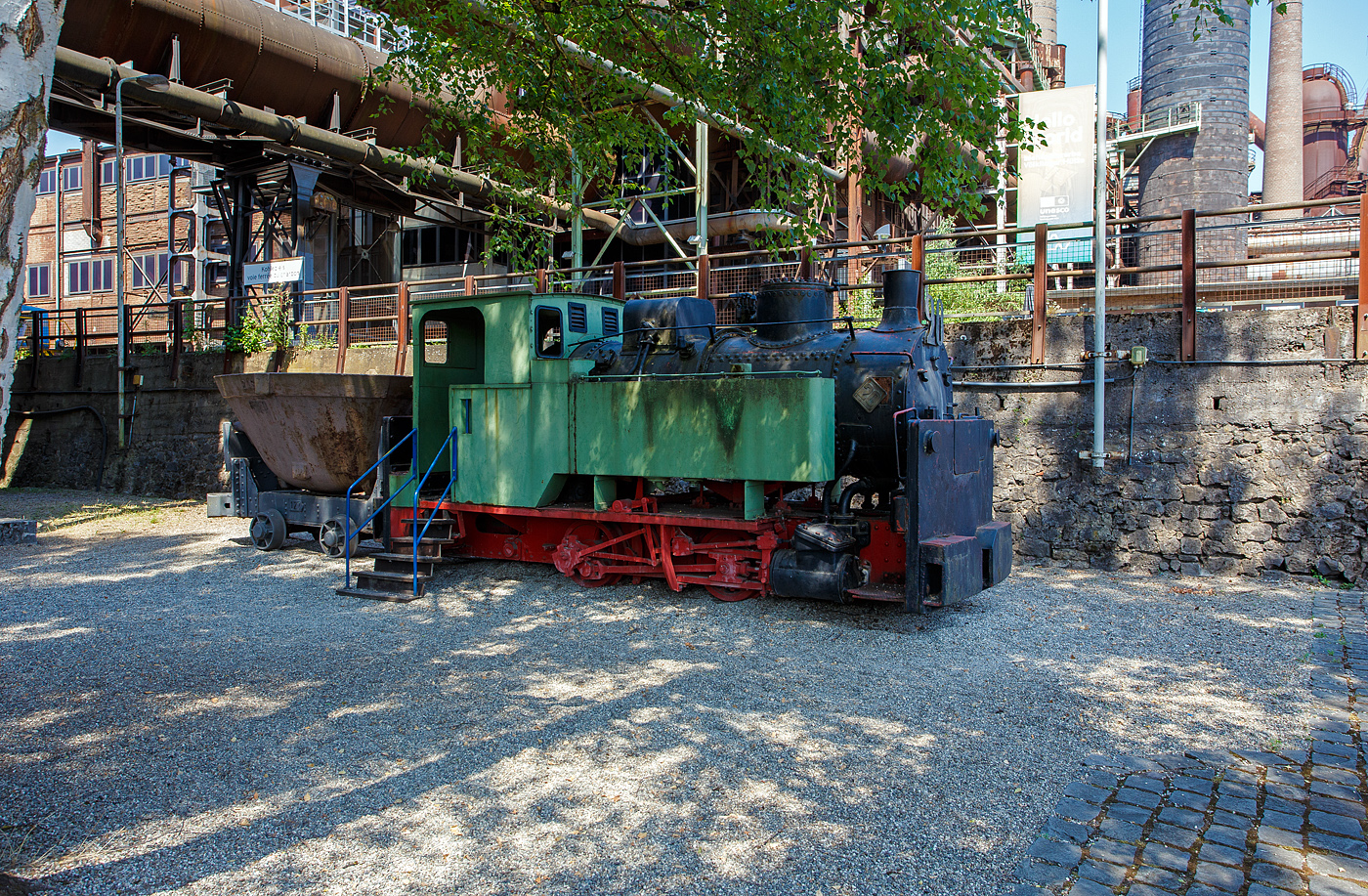 Die 785 mm Schmalspur-Dampflokomotive Nr. 20 der Völklinger Hütte (ehem. Röchling'schen Eisen- und Stahlwerke GmbH in Völklingen/Saar, ab 1982 ARBED-Saarstahl GmbH, ab 1986 Saarstahl Völklingen GmbH) hier am 01 Juli 2025 abgestellt, mit dem Schlackewagen 127 (mit Kübel ARBED 219), als Denkmal beim Biergarten Völklinger Hütte. Heutiger Eigentümer ist die Initiative Völklinger Hütte e.V..

Die Dampflok vom Typ Krauss XXXII kk wurde 1904 von Krauss & Comp. in München unter der Fabriknummer 5168 gebaut und an die RESW - Röchling'sche Eisen- und Stahlwerke AG in Völklingen geliefert und dort als Lok  21  in den Bestand eingereiht. Später tauschte sie mit ihrer Schwesterlok 20 die Betriebsnummer und wurde fortan als Lok  20  eingesetzt. 1978 erfolgte die Aufstellung auf einem Spielplatz in Völklingen-Ludweiler. Im Oktober 1990 übernahm Carreau Wendel vom Musée du bassin houllier Lorrain im französischen Moselle die Lok als Ausstellungsstück. Von dort konnte sie 1997 nach Völklingen zurück und nach einer äußerlichen Aufarbeitung 1998 von der Initiative Völklinger Hütte e.V. in der Alten Völklinger Hütte aufgestellt werden.

Auf dem ehemals 20,4 km langen Schmalspurnetz der Hütte (Spurweite 785 mm) verrichteten bis zu 17 Dampflokomotiven ihren Dienst. Auf der Stahlwerksseite transportierten sie die Schlacke zur Thomasmühle. Die Unterführungen 2 und 3 dienten dem Transport von Stahlblöcken und allgemeinen Gütern zur Versorgung des Walzwerks auf der Eisenwerkseite und dem Abtransport der Walzprodukte. Im Hochofenbereich wurde die Abfuhr der Hochofenschlacke zur Schlackenbrechanlage auf der anderen Saarseite durchgeführt.

Wegen des neuen Stahlwerks, das im Dezember 1980 in Betrieb ging, waren umfangreiche Umbaumaßnahmen notwendig. Der Roheisenkanal wurde tiefer gelegt, die Verbindung zum alten Stahlwerk unterbrochen und im Bereich des Hochofens 6 musste ein neuer Gleisanschluss hergestellt werden. Dadurch musste für die schmalspurige Schlackenbahn eine Hubbrücke gebaut werden, da sich Schmalspurgleis und Roheisengleis auf unterschiedlichem Niveau befinden.

Die baugleiche Lokomotive 21 ist auch vorhanden, sie steht mit zwei Schlackenwagen am Ausgang des Schlackenkanals auf der abgesenkten Hubbrücke in Richtung Schlackenbrechanlage. Leider konnte ich diese nicht entdecken.

TECHNISCHE DATEN der Lok 20 und 21 
Hersteller: Krauss & Comp., München
Typ: XXXII kk
Baujahr: 1904
Spurweite: 785 mm (Schmalspur)
Achsfolge: B
Länge: 5.400 mm
Achsabstand: 1.400 mm
Triebraddurchmesser: 680 mm (neu)
Breite: 1.800 mm
Höhe: 2.650 mm
Dienstgewicht: 16 t
Höchstgeschwindigkeit: k. A. 
Leistung: 80 PS
Zugkraft: 3 t
Zylinder: 2 Stück, Ø 260 mm x 380 mm Hub
Dampfdruck: 14 bar
Heizfläche: 20,62 m²
Rostfläche: 0,43 m“
Anzahl Siederohre: 80 Stück
Durchmesser Siederohre: 39/42 mm
Länge Siederohre: 2.510 mm
Wasservorrat: 1.400 l (1,4 m³)
Kohlenvorrat: 300 kg