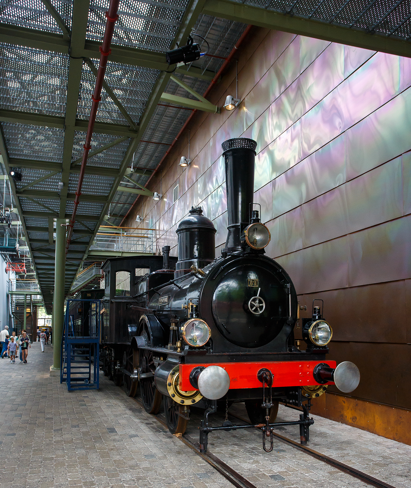 Die Dampflokomotive HSM 89  Nestor , sp�ter NS № 1010 (NS-Baureihe 1000) am 24 Juni 2025 im Het Spoorwegmuseum (bis 2005 NSM - Nederlands Spoorwegmuseum / Niederl�ndischen Eisenbahnmuseum) in Utrecht im ehemaligen Bahnhof Maliebaan.   

Die Dampflok wurde 1879 von Borsig in Berlijn-Tegel unter der Fabriknummer 3730 gebaut und an die damalige HSM - Hollandsche IJzeren Spoorweg-Maatschappij (Holl�ndische Eisenbahn-Gesellschaft) geliefert, 1880 erfolgte die Inbetriebnahme.

HSM 89  Nestor  ist eine niederl�ndische 1B (2-4-0) Tenderlokomotive, die vom Niederl�ndischen Nationalbahnmuseum erhalten wurde. 29 seiner Art wurden f�r den NS-Vorg�nger Hollandsche IJzeren Spoorwegmaatschappij (HSM) gebaut.

In den fr�hen Tagen war die HSM die einzige Eisenbahngesellschaft in den Niederlanden, die ihren Lokomotiven immer einen Namen sowie eine Nummer gab. Der Nestor zum Beispiel wurde auch die Zahl 89 zugewiesen. Trotz der Tatsache, dass es immer als  Der Nestor  bezeichnet wurde, zeigt das Namensschild nur  Nestor . Diese Serie von Lokomotiven wurde  Tenners  genannt, weil sie mit Dampfdruck in H�he von 10 Atmosph�ren umgehen konnten. Ein bemerkenswertes Merkmal dieser Lokomotivserie ist, dass der F�hrerstand der aus Holz und nicht aus Metall besteht. Ihre Lokf�hrer und Heizer wurden daher manchmal  Woonwagenbewoners  genannt, was ungef�hr  Caravan-Bewohner  bedeutet. Metallstangen, die als Schienenw�chter bekannt sind, wurden an der Vorderseite der Rahmenplatten befestigt. Der Lokf�hrer konnte Besen zu diesen Wachen montieren, um die Schienen unter verschneiten Bedingungen zu r�umen. Wie alle Lokomotiven hat der Nestor auch Fettt�pfe montiert. Diese T�pfe w�ren mit Fett und Garn gef�llt oder gef�hlt, um die Achsen zu schmieren.

HSM
Mit dem Ausbau einer Eisenbahnlinie und der Er�ffnung einer weiteren 1879 wurde die Notwendigkeit, ihre Sammlung von Lokomotiven zu erweitern, deutlich und bestellte 29 dieser Maschinen in Borsig. Sie waren 74-82, 89-98 und 116-125 nummeriert, weil die HSM Lokomotiven anhand des Empfangsauftrags nummerierte. Die Serie hie� EOS - JORDAENS, die Namen von 74 und 125. Trotz der Namensschilder, die nur einen Namen haben, wurden sie in der Sprache immer als Der Name bezeichnet (z. B.  Der Nestor  statt  Nestor ).

Bei ihrer Ankunft erhielten verschiedene �ltere Lokomotiven weniger wichtige Arbeiten, die noch �ltere Lokomotiven aus dem Dienst zogen. Mit der Ankunft neuer Sharp Stewart-Lokomotiven im Jahr 1890 wurden die EOS - JORDAENS auf sekund�re, weniger wichtige Aufgaben reduziert, wie sie es zuvor bei �lteren Lokomotiven getan hatten. Sie arbeiteten in den Depots Almelo, Amsterdamer Hauptbahnhof, Apeldoorn, Haarlem und Zutphen und den meisten gro�en Stationen entlang einer Linie nach Osten nach Westen mitten im Land. Sp�ter verteilten sie sich noch mehr auf andere HSM-Depots, die immer mehr auf Rangierarbeiten reduziert wurden.

NS
Bedingt durch den Ersten Weltkrieg wurde 1917 eine Zweckgemeinschaft der unabh�ngigen Eisenbahngesellschaften HSM beziehungsweise HIJSM (Hollandsche IJzeren Spoorweg-Maatschappij) und SS (Maatschappij tot Exploitatie van Staatsspoorwegen) gegr�ndet. Die beiden Gesellschaften errichteten einen gemeinsamen Bahnbetrieb, blieben aber rechtlich unabh�ngig. Dies �nderte sich 1938, als HIJSM und SS vollst�ndig fusionierten und in der AG NS (Nederlandse Spoorwegen, fr�her Nederlandsche Spoorwegen geschrieben) aufgingen. Das Rollmaterial wurde bereits 1921 zusammengelegt, so wurden die EOS - JORDAENS zu NS 1001 - 1029 nummeriert. Die NS vergr��erte auch die Fenster in der Kabine.

Bis 1923 befanden sich acht von ihnen am Hauptbahnhof Utrecht, der vor der Fusion ausschlie�lich Staatsspoorwegen-Gebiet war. Sie wurden ausschlie�lich zum Rangieren verwendet. In Amersfoort waren f�nf weitere mit der gleichen Arbeit und in Winterswijk, vier, obwohl diese vier auch Z�ge zu einigen nahe gelegenen Bahnh�fen fuhren. 1924 wurde jedoch die Mehrheit der NS-1000 in die westlichsten Depots, einschlie�lich Apeldoorn, verlegt.

Elf der Baureihe wurden 1921-1923 ausgemustert. Der NESTOR �berlebte mit dem Rest bis 1934. In diesem Jahr schickte NS alle bis auf zwei, NS 1010 & 1011, auf den Schrottplatz. Ein Jahr sp�ter folgte auch NS 1011. Obwohl der NESTOR (NS 1010) auch 1934 abgeschafft werden sollte, half die NVBS (Niederl�ndische Vereinigung der Interessenten in der Eisenbahn- und Stra�enbahnindustrie) dabei, sie f�r das noch zu bauende niederl�ndische Eisenbahnmuseum zu erhalten, so dass sie in Boxtel gelagert wurde.

Die Lokomotive wurde zusammen mit anderen f�r das Museum reservierten Rollmaterial ein paar Mal w�hrend des Zweiten Weltkriegs bewegt. 1944 war die NESTOR in Maastricht, dort wurde sie den zur�ckweichenden Deutschen schwer besch�digt (gesprengt). Als der Krieg endete, wurde die Maschine eine Weile in Almelo gelagert, immer noch schwer besch�digt. Erst 1953 erteilte die NS die Erlaubnis, sie zu reparieren. Die Reparatur wurde 1964 abgeschlossen. In diesem Jahr fuhr die Lokomotive die letzten 100 Meter alleine zum niederl�ndischen Eisenbahnmuseum, obwohl die n�tige Energie mit Druckluft aus einem Diesel geliefert wurde.

Der NESTOR war die letzte NS-Lokomotive, die f�r Museen restauriert wurde. Sie bekam ihren Look aus den 1880er Jahren, einschlie�lich der Holzkabine mit hohen Fenstern.

Im niederl�ndischen Eisenbahnmuseum ist NESTOR nicht der einzige Ex-NS 1010. Die Baunummer der Baureihe 1000 wurde sp�ter f�r Elektrolokomotiven verwendet, deren Zehnter ebenfalls erhalten blieb.

TECHNISCHE DATEN:
Typ: Schnelldampflok 
HSM-Serie: EOS - JORDAENS, ab 1921 NS 1001 - 1029
Spurweite: 1.435 mm (Normalspur)
Achsfolge: 1B (engl. 2-4-0)
L�nge �ber Puffer: 14.160 mm (Lok 8.260 mm + Tender 5.900 mm)
Treibraddurchmesser: 1.860 mm (neu)
H�chstgeschwindigkeit: 90 km/h
Gesamtheizfl�che: 93,5 m�
max. Dampfdruck: 10,3 kg/cm�
Anzahl der Zylinder: 2 (� 406 mm x 558 mm Hub)
Eigengewicht: Lok 38,8 und Tender 25,1 t = 63,9 t
Anh�ngelast: 3560 kg
Wasservorrat: 9 m�
Kohlevorrat: 3,6 t
