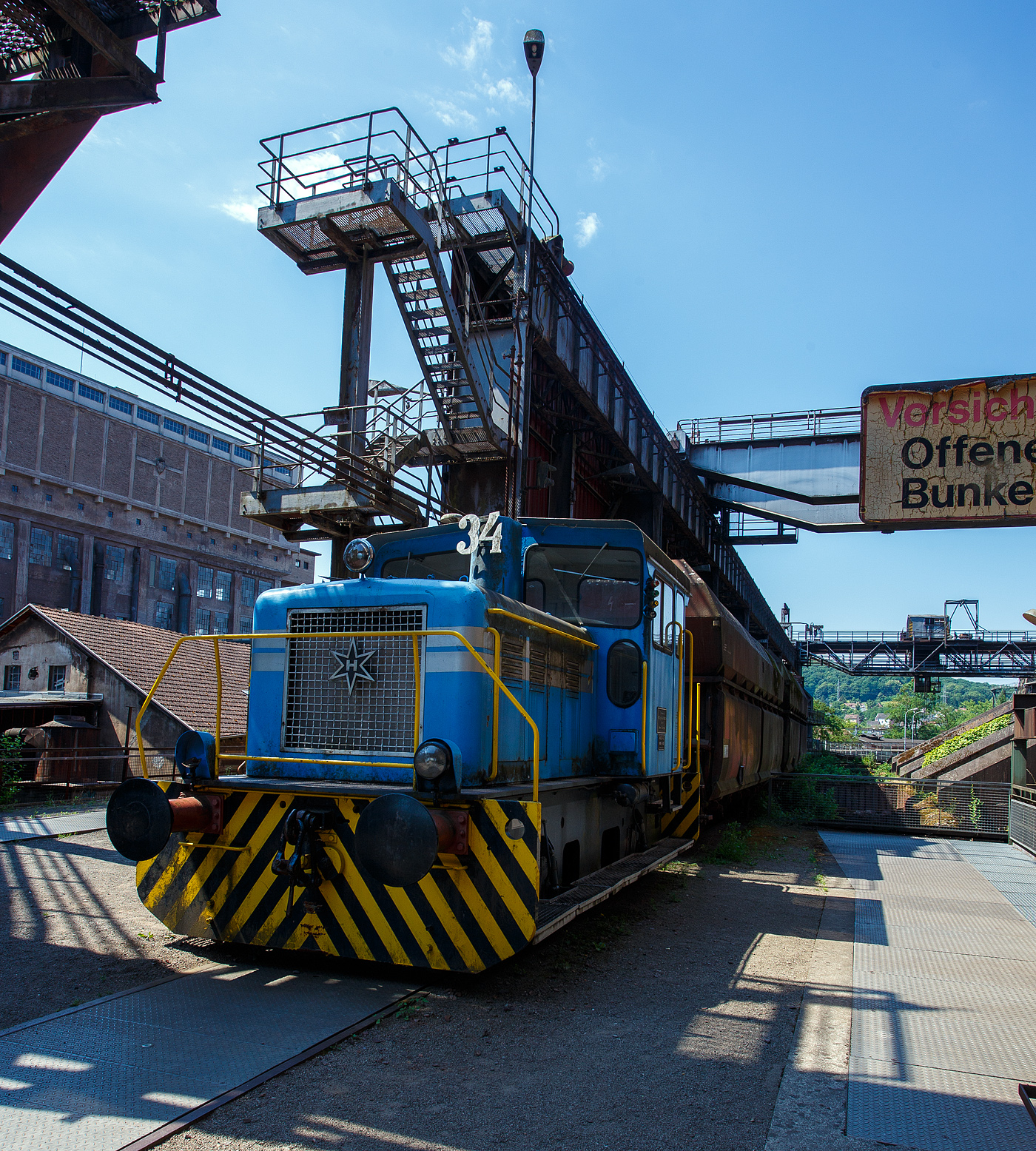 Die Dieselhydraulische Normalspur-Lokomotive Nr. 34, eine Henschel DHG 240 B  Esslinger , der Völklinger Hütte (ehem. Röchling'schen Eisen- und Stahlwerke GmbH in Völklingen/Saar, ab 1982 ARBED-Saarstahl GmbH, ab 1986 Saarstahl Völklingen GmbH) hier am 01 Juli 2025 abgestellt als Denkmal, mit zwei selbstentladende Schüttgutwagen der Gattung Fad oder Falns am Eingang der Rohstoffentladebunker in Richtung Hüttenbahnhof. Die Darstellung steht für den Rohstofftransport der Hütte zur Herstellung des Sintergutes. Die Lok war auch auf Bundesbahn Gleisen zugelassen. Heutiger Eigentümer der Lok und Wagen ist die Initiative Völklinger Hütte e.V..

Die zweiachsige dieselhydraulische Lok mit Antrieb über Gelenkwellen wurde 1969 von Henschel in Kassel unter der Fabriknummer 31210 gebaut und an die Röchling'sche Eisen- und Stahlwerke GmbH in Völklingen als Lok 34 geliefert. Weiterer Lebenslauf/Umfirmierungen: 
01.01.1971 > Stahlwerke Röchling-Burbach GmbH, Völklingen  34  
Juni 1982 	 > ARBED-Saarstahl GmbH, Völklingen  34  
Juni 1986 	 > Saarstahl Völklingen GmbH, Völklingen  34  
10.05.1989 > Saarstahl AG, Völklingen  34  
Später an MGS - Metallurgische Gesellschaft Saar mbH, Völklingen  34  
März 1999 	an Weltkulturerbe Völklinger Hütte - Europäisches Zentrum für Kunst und Industriekultur GmbH, Völklingen und Aufstellung als Denkmal. 

Die Lokomotive war bis zu einem Motorschaden 1997 in verschiedenen Bereichen eingesetzt: Transport von Duplexeisen zwischen der Mischerhalle und dem Siemens-Martin-Werk, im Bereich des Siemens-Martin-Werks, der Elektroöfen und des Hammerwerks sowie nach Einbau einer Funkfernsteuerung im Hafenbereich der Saarstahl AG.

Mit den Fahrzeugen der Typenreihe DHG 160 B / DHG 200 B / DHG 240 B und DHG 350 B bot Henschel Anfang der 1960er Jahre Gelenkwellenmaschinen für den unteren Leistungsbereich an, deren konstruktiver Ursprung nicht in Kassel, sondern in Esslingen der lag. Im Jahr 1961 übernahm Henschel das Diesellok-Typenprogramm der Maschinenfabrik Esslingen. Es wurde eine bei der Maschinenfabrik Esslingen als Typ Bern in Auftrag gegebene Lokomotive als DHG 325 in Kassel fertig gebaut und an den Auftraggeber, die Duisburger Kupferhütte, ausgeliefert. Der Typ Bern stellte auch die Basis für das oben genannte Typenprogramm dar. Dieses unterschied sich zwar optisch deutlich von seinem Esslinger Ursprung, übernahm aber die wesentlichen konstruktiven Merkmale der Fahrzeuge aus Esslinger Produktion. Gleichzeitig gab es keinen technischen und optischen Bezug zu den bisher im eigenen Henschel-Typenprogramm angebotenen Fahrzeugen.

Bei den neuen Gelekwellenfahrzeugen gab es leistungsmäßig deutliche Überschneidungen mit dem kleinsten Typ der 3. Generation, der stangengetriebenen DH 240 B. Diese wurde dann in der 4. Generation durch die DHG 240 B ersetzt.

Die Henschel DHG 240 B wurde zwischen 1963 und 1971 in 34 Exemplaren gebaut. Die Loks gingen vornehmlich an Chemiebetriebe und Raffinerien in Deutschland und der Schweiz. Von diesen wurden elf Loks explosionsgeschützt ausgerüstet. Einzelne Loks wurden ebenfalls mit Funkfernsteuerung ausgerüstet. Mit weitem Abstand am stückzahlenstärksten ist die DHG 240 B, die im Gegensatz zu ihren schwächeren  Schwestern  nicht mit einem DIWABUS-Getriebe von Henschel, sondern mit einem Voith-Hydraulikgetriebe ausgestattet ist. Zahlreiche Fahrzeuge stehen auch heute noch im täglichen Einsatz.

TECHNISCHE DATEN:
Baujahre: 1963 bis 1971
Gebaute Stückzahl: 34
Spurweite:	1.435 mm (Normalspur)
Achsfolge: B
Länge über Puffer: 7.160 mm
Achsabstand: 2.800 mm
größte Breite: 3.000 mm
größte Höhe über Schienenoberkante: 3.600 mm
Raddurchmesser: 850 mm (neu)
kleinster befahrbarer Gleisbogen: 35 m
Eigengewicht: 28 t
Max. Achslast: 16 t
Dieselmotor: wassergekühlter 6 Zylinder / 4-Takt-Diesel-Reihenmotor mit Turboaufladung, ohne Ladeluftkühlung vom Typ Henschel 6R 1416 A, Hubraum 13,3 l.
Leistung: 177 kW (240 PS)
Drehzahl: 1.600 U/min
Höchstgeschwindigkeit: 30 / 60 km/h
Leistungsübertragung: hydraulisch (Voit-Getriebe)
Kraftstoffvorrat: 400 l
