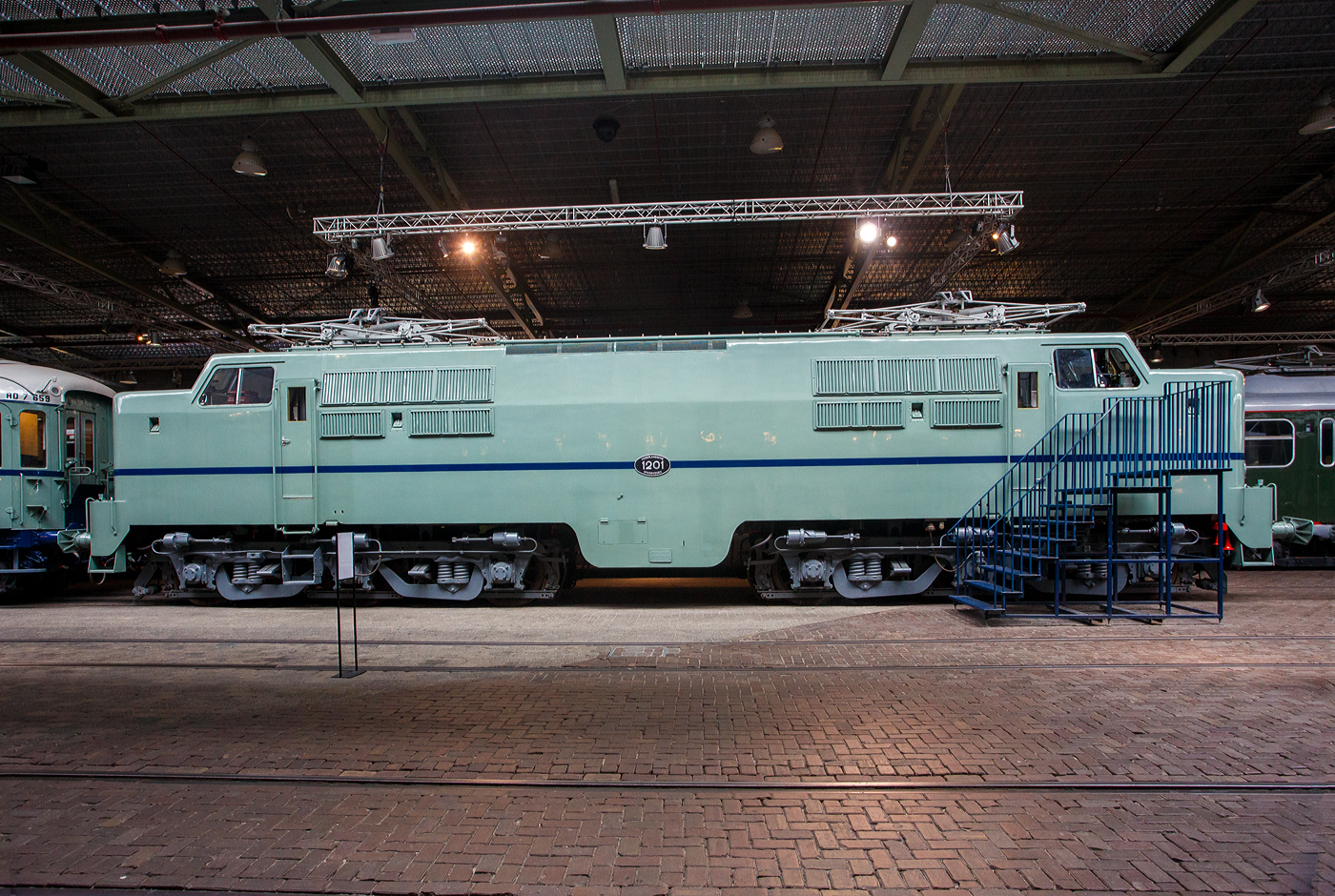 Die Elektrolokomotive NS 1201 der NS-Baureihe 1200 am 24 Juni 2025 im Het Spoorwegmuseum (bis 2005 NSM - Nederlands Spoorwegmuseum / Niederländischen Eisenbahnmuseum) in Utrecht im ehemaligen Bahnhof Maliebaan. Ein Eisenbahnmuseum das zum Verweilen einlädt.

Die Lok wurde 1951 von Werkspoor in Utrecht unter der Fabriknummer 928 gebaut (Baldwin-Lizenz) und an die NS - Nederlandse Spoorwegen (Niederländischen Staatsbahnen, heute Niederländische Eisenbahnen AG) geliefert. Wobei die Entwicklung und Konstruktion, sowie auch einige Teile, von Baldwin Locomotive Works in Philadelphia (USA) stammt. Auch die Entwicklung der Elektrotechnik stammt aus den Vereinigten Staaten und zwar von Westinghouse Electric Corporation (Monroeville), diese wurde von Heemaf in Hengelo (Westinghouse-Lizenz) geliefert.

Die Baureihe 1200 der Niederländischen Eisenbahnen (NS) ist eine sechsachsige Elektrolokomotivbaureihe aus den Jahren 1951 bis 1953. Die Lokomotiven waren bis 1998 bei der NS im Einsatz.

Die Nase war der erste Teil dieser unglaublich starken und robusten Lokomotive, der ins Auge fiel. Sie sollte dem Lokführer bei Kollisionen einen besseren Schutz bieten. Diese Lokomotiven wurden in den Niederlanden nach einem amerikanischen Design und mit einigen amerikanischen Teilen gebaut, die teilweise durch amerikanische Dollar über den Marshall-Plan finanziert wurden. Die NS nutzte diese Lokomotiven, um sowohl Personenzüge als auch Güterzüge zu ziehen. Ihre wichtigste Aufgabe war es Schnellzüge zu ziehen. Als sich die NS 1995 trennte, wurden alle 1200er Jahre an NS Cargo übertragen und begannen im Güterverkehr zu arbeiten. 

Nach Ende des Zweiten Weltkrieges gab es großen Fahrzeugmangel bei der NS. Da man keine Erfahrungen mit dem Bau elektrischer Lokomotiven hatte, kaufte man Lokomotiven bei verschiedenen Lieferanten. Die bei Baldwin konstruierten Lokomotiven wurden bei Werkspoor in Utrecht gefertigt. Die bei Westinghouse entwickelte Elektrotechnik wurde von Heemaf in Hengelo geliefert. Von den ursprünglich 75 georderten Lokomotiven wurden nur 25 ausgeliefert. Der amerikanische Einfluss ist an den hochliegenden Führerständen und den großen, dem Schutz des Lokführers dienenden Vorbauten erkennbar. Am 30. November 1950 wurden 25 Loks bestellt, am 27. September 1951 konnte die erste Probefahrt stattfinden. Am 9. Februar 1952 wurde die 1201 offiziell in Dienst gestellt, der Preis pro Lok betrug 765.000 NFL (ca. 347.000 Euro).

Die Loks hatten sechs Motoren des Typs TM94. Die ersten 14 Lokomotiven waren türkis lackiert, danach setzte sich wegen des sichtbaren Abriebs der Stromabnehmer eine rotbraune Farbgebung durch. Ab 1954 erhielten alle Loks eine blaue Farbgebung, 1971 erhielt die erste Lok die neuen NS-Hausfarben grau/gelb, 1983 war auch die letzte Lok entsprechend umlackiert. Am 30. März 1998 endete der planmäßige Einsatz bei der NS.

Fünf weitere Lokomotiven (1214, 1215, 1218, 1221, 1225) wurden 1999 an die ACTS Nederland BV verkauft. Diese vermietete 2010 drei Lokomotiven (1214, 1215, 1225) an die Eisenbahngesellschaft Euro-Express Treincharter (EETC). Von der wurden sie bis April 2015 unter anderem vor den Nachtzügen Niederlande–Österreich zwischen Amsterdam und Venlo eingesetzt. Fünf Maschinen (1201, 1202, 1211, 1218, 1221) sind als Museumslokomotiven erhalten. NS 1211 ist Botschafterlok im Bahnpark Augsburg. Leihgeber ist das Eisenbahnmuseum Utrecht.

TECHNISCHE DATEN:
Hersteller mech.: Werkspoor (in Lizenz von Baldwin)
Hersteller Elektrik: Heemaf (in Lizenz von Westinghouse)
Baujahre: 1950 bis 1953
Ausmusterung bei NS: bis 1998 
Gebaute Anzahl: 25
Nummerierung: NS 1201 bis 1225
Spurweite: 1.435 mm (Normalspur)
Achsfolge: Co’Co’ 
Länge über Puffer: 18.086 mm
Höhe: 4.064 mm
Treibraddurchmesser: 1.100 mm (neu)
Dienstgewicht: 108 t
Antriebsmotoren: 6 Stück Heemaf vom Typ TM 94
Getriebeübersetzung: 20 : 71
Höchstgeschwindigkeit: 130 km/h (Planmäßig) / 150 km/h (technisch möglich)
Stundenleistung: 6 x 392 kW = 2.350 kW
Dauerleistung: 6 x 367 kW = 2.200 kW
Anfahrzugkraft: 195 kN
Dauerzugkraft: 164 kN bei 80 km/h / 64,7 kN bei 123 km/h
Stromsystem: 1,5 kV DC (=)
Kleister befahrbarer Gleisbogen: R 100 m