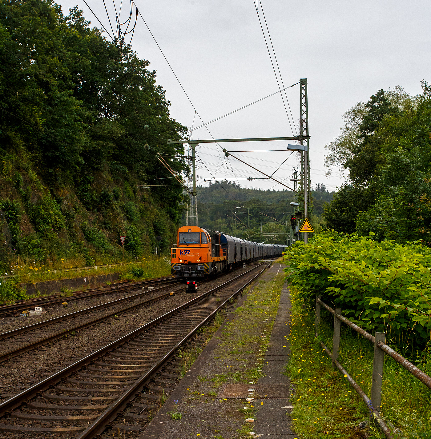 Die KSW 43 (92 80 1273 018-2 D-KSW), die asymmetrische Vossloh G 2000 BB der KSW (Kreisbahn Siegen-Wittgenstein) fährt am 03 Juli 2024, mit einem beladen Coilzug (16 Stück VTG Coilwagen der Gattung Shimmns-ttu, VTG Bez. F46.520D) vom Hafen Duisburg kommend, durch den Bahnhof Scheuerfeld(Sieg) in Richtung Herdorf via Betzdorf. 

In Betzdorf (Sieg) geht es dann über KBS 462 „Hellertalbahn“ nach Herdorf zum Rbf der KSW (Betriebsstätte FGE -Freien Grunder Eisenbahn). Für die Weiterfahrt auf den Pfannenberg (in Neunkirchen-Salchendorf) über nimmt dort dann die KSW 47 (92 80 1271 027-5 D-KSW), ex D 2 der HFM, eine Vossloh G 1000 BB, den Zug.
