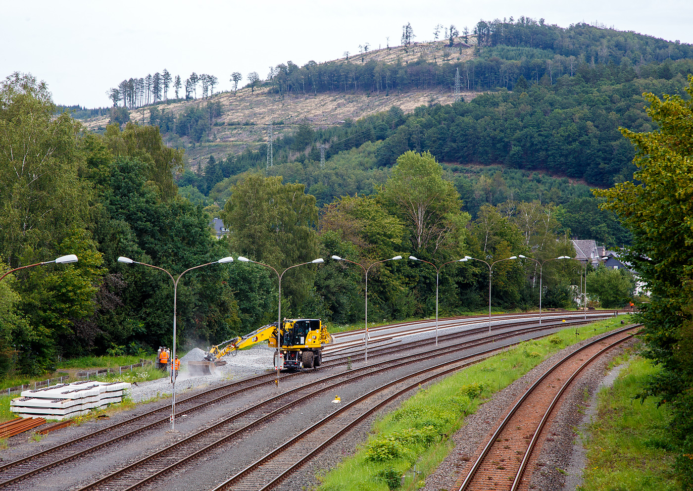 Die KSW (Kreisbahn Siegen-Wittgenstein) lässt, auf ihren Rangierbahnhof in Herdorf der Betriebsstätte FGE -Freien Grunder Eisenbahn (KSW NE447 / DB-Nr. 9275), nach und nach die Gleise erneuern. 
Hier ist am 24.08.2023, der Caterpillar CAT M323F Zweiwegebagger mit hydrostatischem Schienenradantrieb (Serien-Nr. RH600 189), Kleinwagen Nr. D-ZBM 99 80 9902 521-0 der ZEPPELIN Baumaschinen GmbH (Garching), ein Mietbagger vermietet an die Firma  W. Hundhausen Bauunternehmung GmbH (Siegen), mit dem Einschottern eines Gleises beschäftigt.

Rechts die DB Strecke KBS 462 „Hellertalbahn“. 
