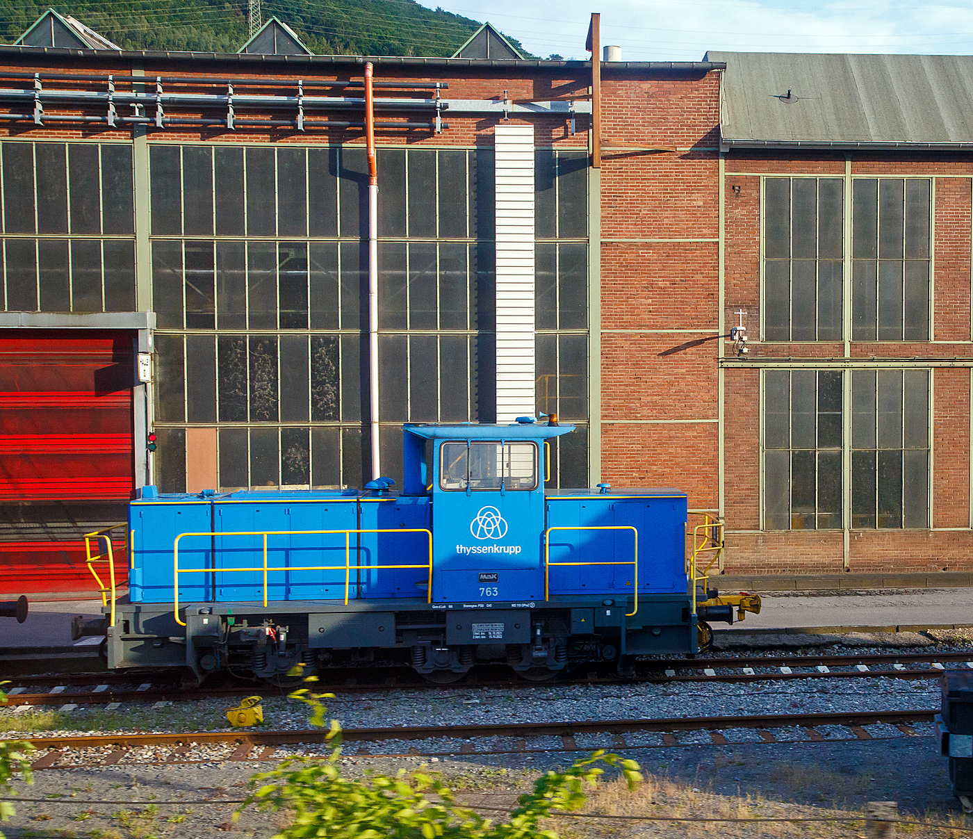 Die Lok 763 der thyssenkrupp Hohenlimburg GmbH, ex 763 Hoesch Hohenlimburg GmbH, ex SK 32 (Siegener Kreisbahn GmbH), am 31.05.2022 auf dem Werksgelände in Hagen-Hohenlimburg, aus einem fahrenden Zug heraus fotografiert.

Die Lok ist eine MaK G 763 C, sie wurde 1990 von der Maschinenbau Kiel GmbH (MaK) unter der Fabriknummer 700099 gebaut und an die damalige Siegener Kreisbahn GmbH als SK 32 geliefert. Zum 23.12.2004 nach der Umbenennung zur Kreisbahn Siegen-Wittgenstein GmbH wurde sie zur KSW 32 und war von 2005 bis 2007 an die MEG - Märkische Eisenbahn-Gesellschaft mbH in Lüdenscheid vermietet. Im Dezember 2007 ging sie über Vossloh Locomotives an die Hoesch Hohenlimburg GmbH als Lok 763, zum 01.07.2016 wurde Hoesch von thyssenkrupp übernommen.