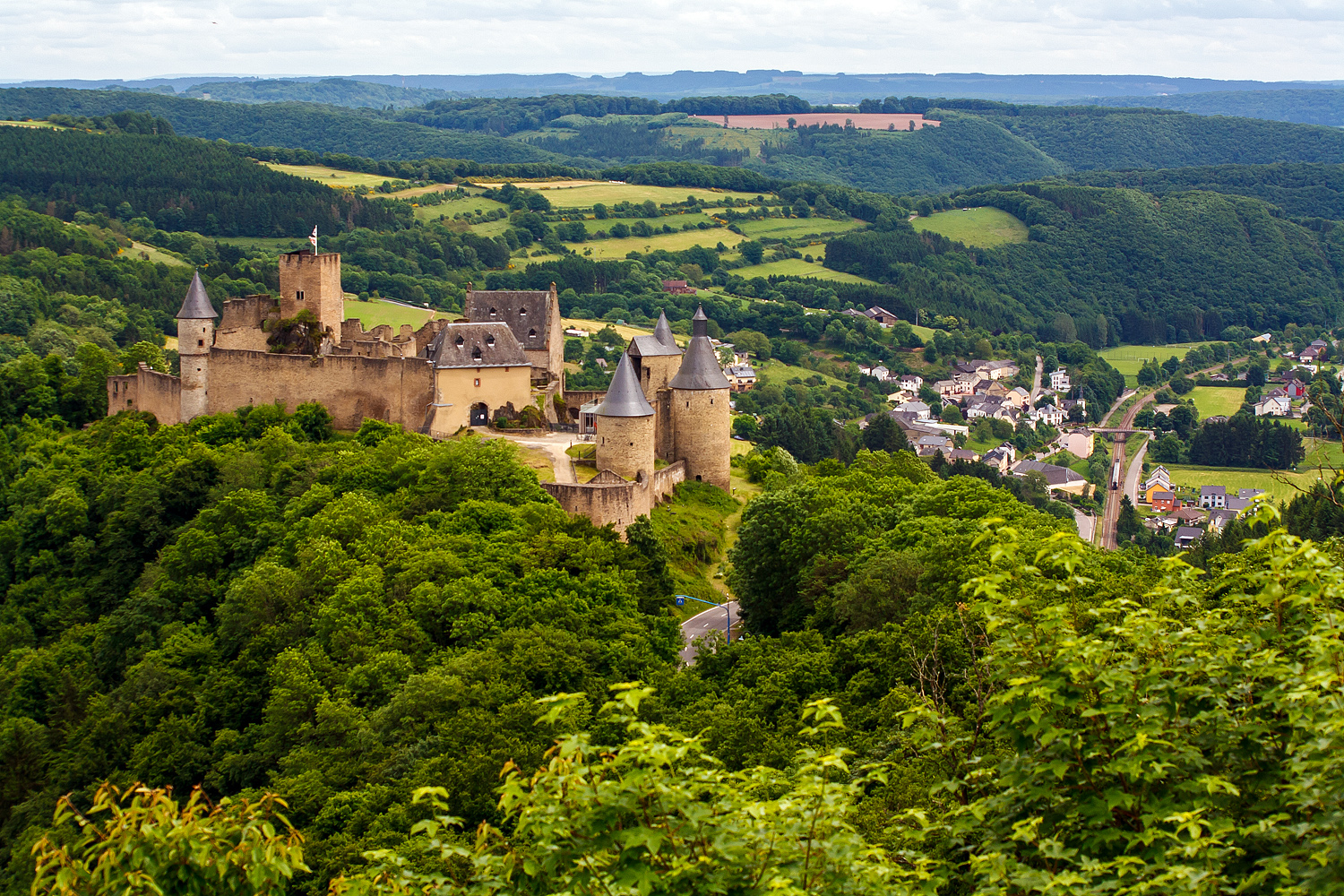 Ein BahnSuchBild: Blick auf die Burg Bourscheid (Burscheid) rechts unten der zu Burscheid geh�rende Ort Michelau am 15. Juni 2013. Eine CFL 3000er, die CFL 3006, f�hrt mit 3 Personenwagen aus Richtung Kautenbach kommend durch Michelau in Richtung Ettelbr�ck bzw. Luxemburg.

Burg Bourscheid ist Luxemburgs gr��te Burg und ein beeindruckendes Bauwerk mit charakteristischen Rundt�rmen, gelegen auf einem Felssporn 150 m �ber der Sauer. Die Burg wurde wahrscheinlich im 10. Jahrhundert erbaut, im 14. und 15. Jahrhundert vergr��ert und erhielt dann die von sechs gotischen T�rmen flankierte Festungsmauer. 