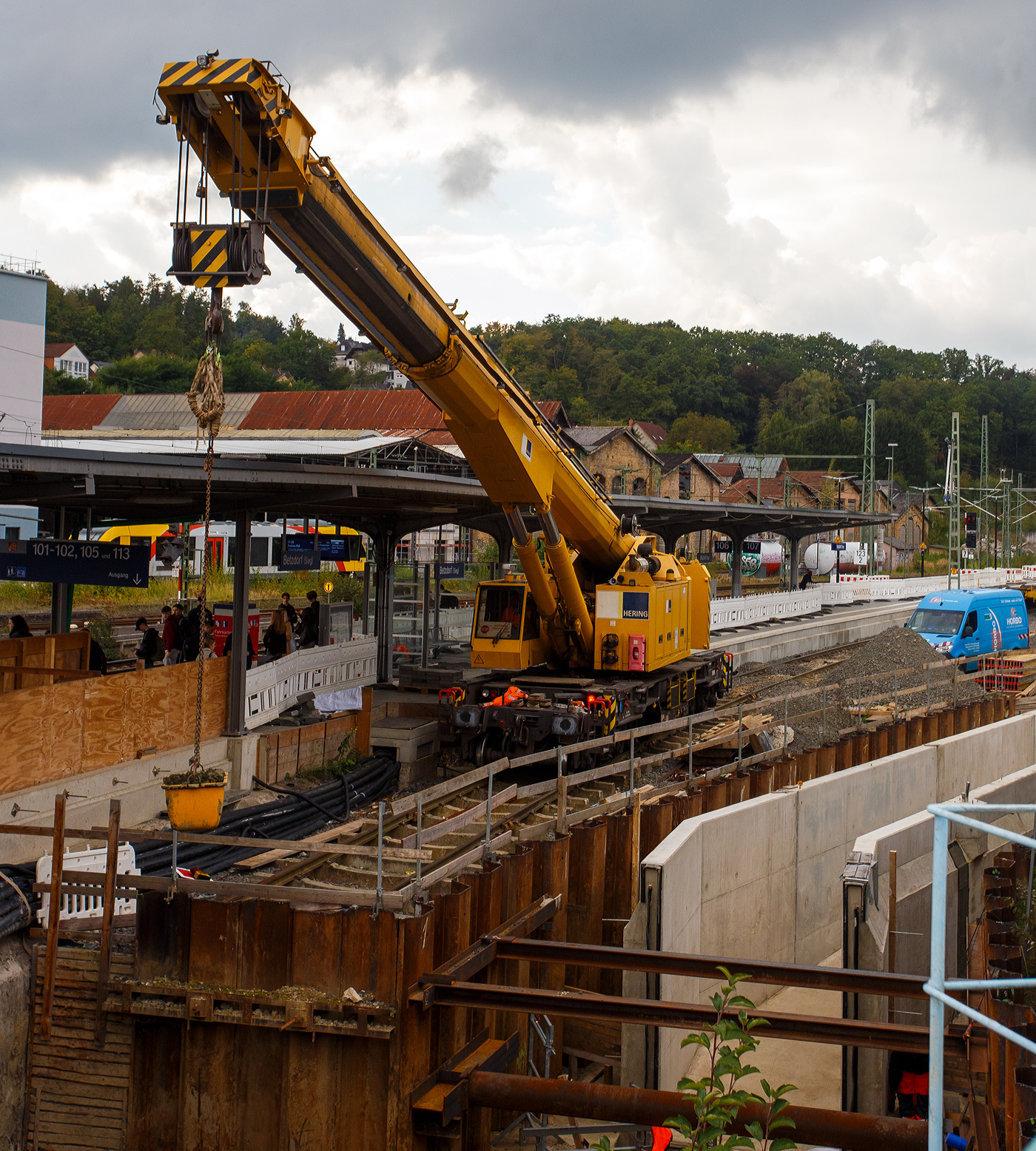 Nun im Einsatz der 125 t KIROW Gleisbauschienenkran KIROW Multi Tasker KRC 810 T (mit Schwerlastausrüstung), Schweres Nebenfahrzeug Nr. D-HGUI 99 80 9419 024-1, ex 97 82 53 513 19-3, der HERING Bahnbau GmbH (Burbach) ist am 29 September 2025 im Betzdorf/Sieg. 

Hier im Bereich unter dem P+R Parkhaus bis zum Fluss Sieg werden Horizontalbohrungen gemacht, vermutlich für erforderliche Entwässerungs-Leitungen. So mussten Bohrrohre in die Baugrube gehoben werden und wie hier der Abraum aus der Baugrube gehoben werden. Eigentlich ist ein 125 t Kran damit unterfordert (überdimensioniert), aber man muss bedenken es wurde eine weite Auskragung (Ausladung vor Puffer) und das freistehend (Nichtabgestützt) benötigt. 

Der hier mit der Ausladung vor Puffer von über 15 m könnte der Kran freistehend 30 t heben, aber darf dann nicht schwenken. Wenn geschenkt werden muss liegt diese dann nur bei 1,5 t. Entscheidend für die Ausladung des Auslegers ist aber auch die hintere Ausladung des Gegengewichts. 